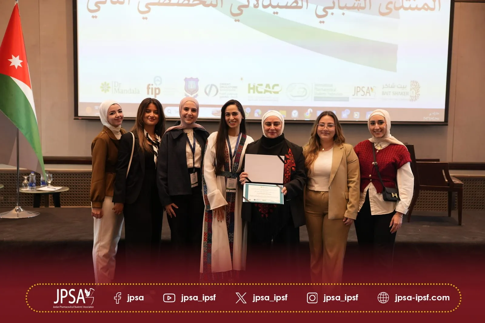A group posing with a certificate beside the Jordanian flag