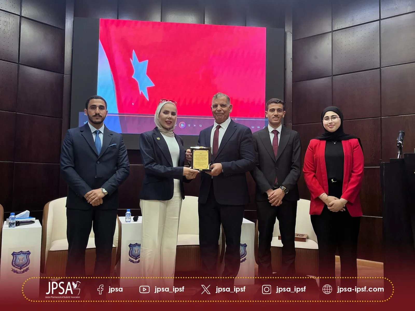 A plaque presented on stage, in front of a screen showing the Jordanian flag
