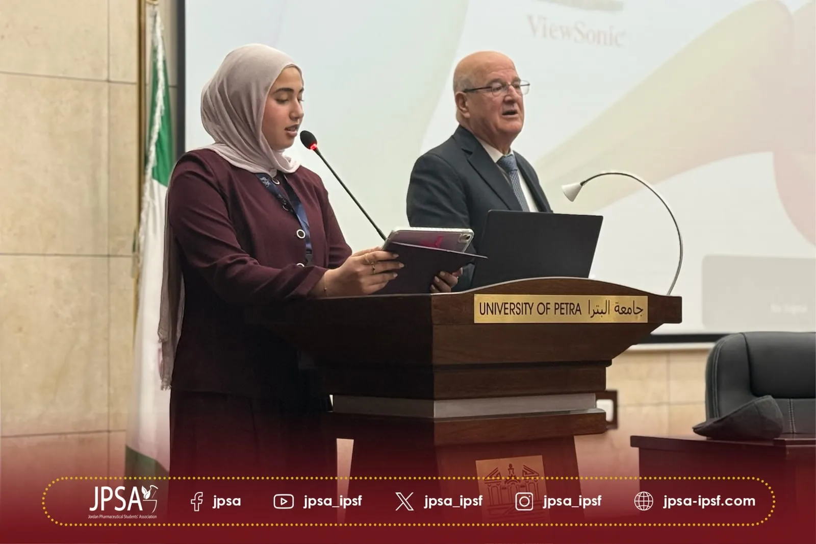 Two people at a lectern, one speaking while reading from a tablet